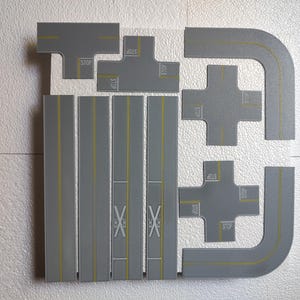May include: A collection of gray toy road pieces, including straight sections, curves, intersections, and a T-intersection. Yellow lines mark the road lanes, and some pieces have "STOP" markings. The set includes railroad crossing symbols.