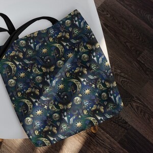 May include: A tote bag with a celestial pattern featuring moons, stars, and other mystical symbols in gold and white against a dark blue and green background. The bag has black handles and is resting on a white chair.