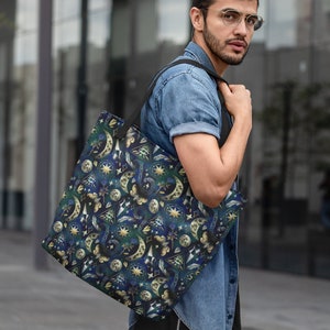 May include: A dark blue tote bag with a celestial pattern of moons, stars, and other mystical symbols in gold and white. The bag has a black strap and is carried over the shoulder.