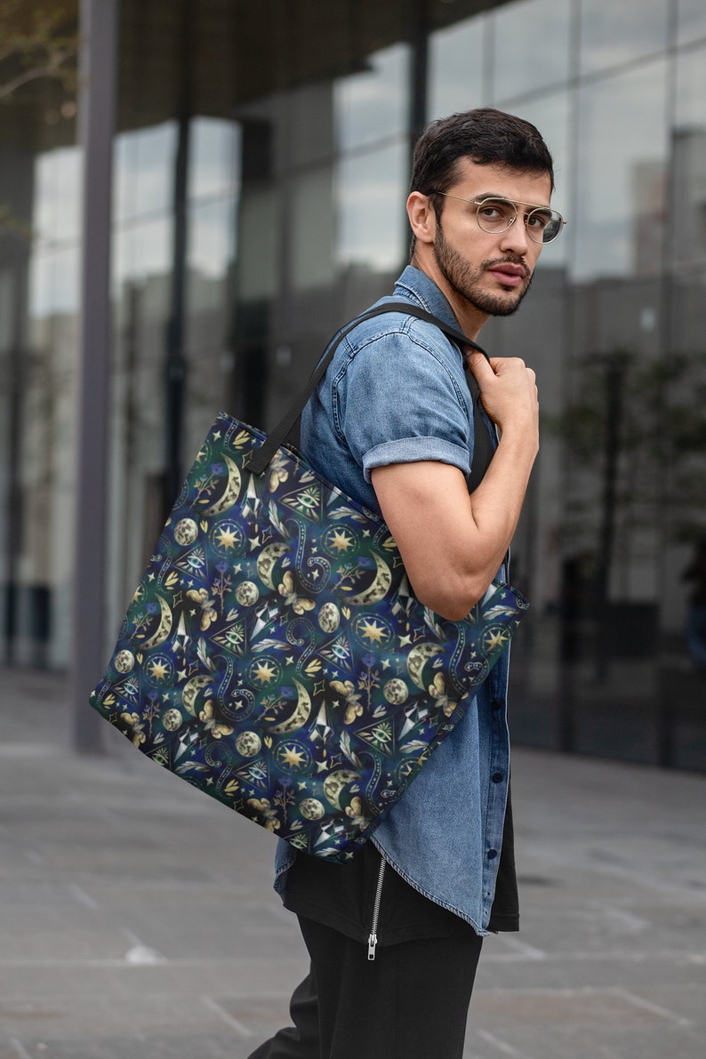 May include: A dark blue tote bag with a celestial pattern of moons, stars, and other mystical symbols in gold and white. The bag has a black strap and is carried over the shoulder.
