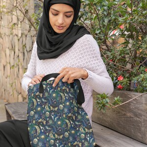 May include: A tote bag with a celestial pattern featuring moons, stars, and other mystical symbols in shades of blue, green, and gold. The bag has black handles and is held by a person wearing a black head covering.