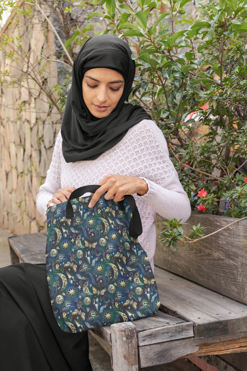 May include: A tote bag with a celestial pattern featuring moons, stars, and other mystical symbols in shades of blue, green, and gold. The bag has black handles and is held by a person wearing a black head covering.