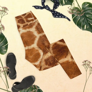 May include: High-waisted leggings with a giraffe print in shades of brown and cream. The leggings are paired with black sandals, a navy blue polka dot scarf, and tropical foliage on a cream background. A stylish and comfortable choice.