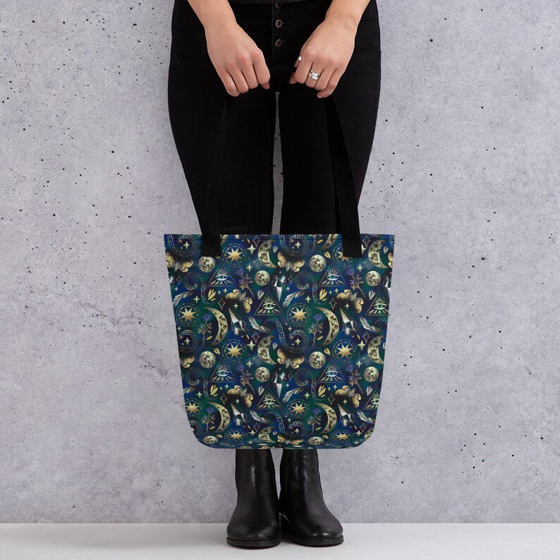 May include: A tote bag with a dark blue background and a repeating pattern of moons, stars, butterflies, and other celestial and occult symbols in gold and white. The bag has black handles.