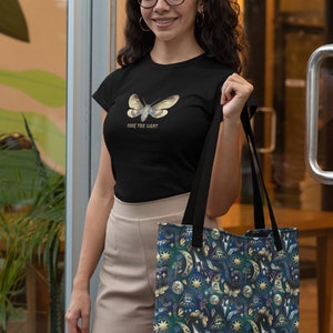 May include: Black t-shirt with a moth graphic and the text "Seek the Light". The person is holding a tote bag with a dark blue celestial pattern featuring moons, stars, and eyes.