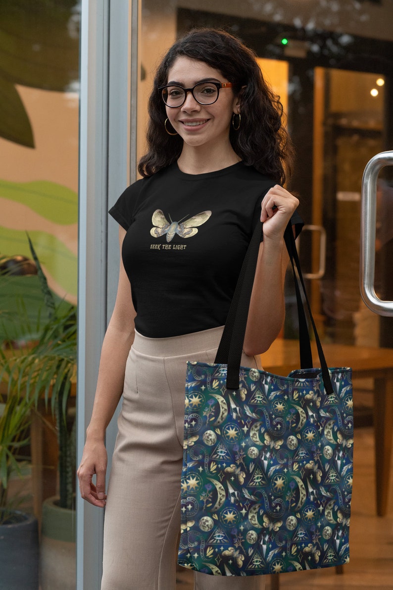 May include: Black t-shirt with a moth graphic and the text "Seek the Light". The person is holding a tote bag with a dark blue celestial pattern featuring moons, stars, and eyes.