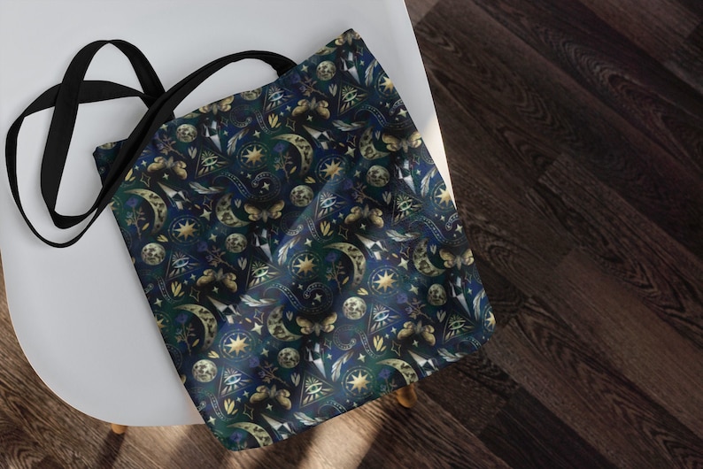 May include: A tote bag with a celestial pattern featuring moons, stars, and other mystical symbols in gold and white against a dark blue and green background. The bag has black handles and is resting on a white chair.