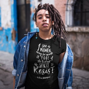 Op de afbeelding: Zwart T-shirt met de quote "I see a women may be made a fool if she had not a spirit to resist" van William Shakespeare in witte letters. De persoon draagt een spijkerjasje.