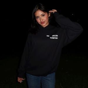 Puede incluir: Una sudadera con capucha negra con el texto blanco "WE BELONG TOGETHER". La sudadera se combina con unos vaqueros azules. La imagen está tomada de noche.