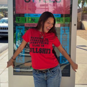 Könnte beinhalten: Ein leuchtend rotes Kurzarm-T-Shirt mit dem schwarzen Text DANCING IS EVERYTHING EVERYTHING ELSE IS BULLSHIT. Das Oberteil wird mit einer blauen Jeans kombiniert, die von einem schwarzen Gürtel mit silberner Schnalle gehalten wird.