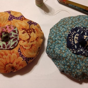 Puede incluir: Dos almohadillas redondas de tela con patrones florales. Una es naranja, morada y verde con un centro rosa y verde. La otra es azul y blanca con un centro azul marino.