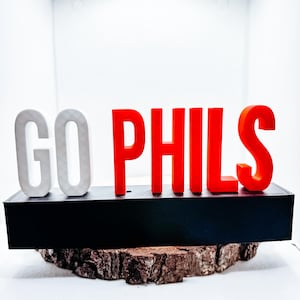 May include: A decorative sign with the words "GO PHILS" in white and red block letters. The letters sit on a black base, which is placed on a piece of wood. The background is white.