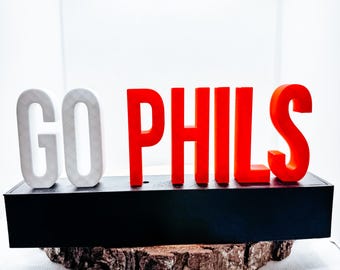 May include: A decorative sign with the words "GO PHILS" in white and red block letters. The letters sit on a black base, which is placed on a piece of wood. The background is white.