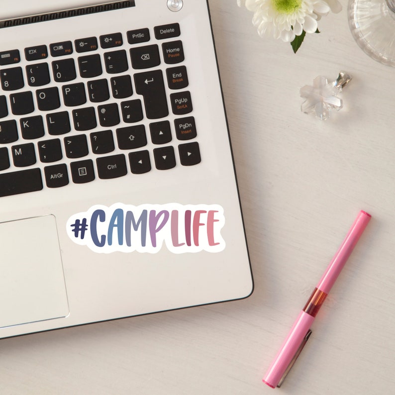 May include: A laptop computer with a sticker that says "#CAMPLIFE" in a rainbow gradient. The sticker is on the bottom right corner of the laptop. A pink pen is on the table next to the laptop.
