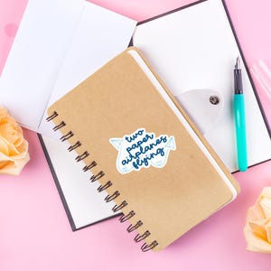 以下が含まれることがあります： 白いステッカーに「two paper airplanes flying」と書かれた茶色のノート。ステッカーにはハートのついた紙飛行機が2機描かれている。ノートは開いていて、上に白い紙が置かれている。ノートの右側には青いペンと白い紙がある。