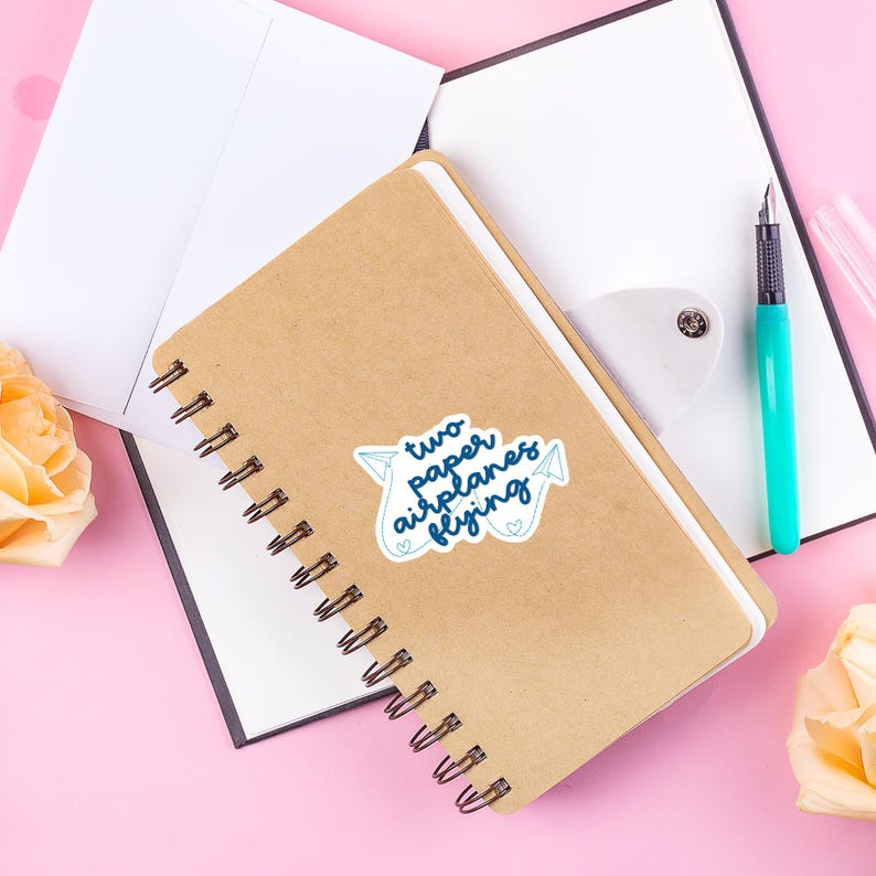 以下が含まれることがあります： 白いステッカーに「two paper airplanes flying」と書かれた茶色のノート。ステッカーにはハートのついた紙飛行機が2機描かれている。ノートは開いていて、上に白い紙が置かれている。ノートの右側には青いペンと白い紙がある。