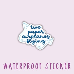 以下が含まれることがあります： 防水ステッカー。青いテキストで「two paper airplanes flying」と書いてあり、青い紙飛行機のアウトラインが2つあります。