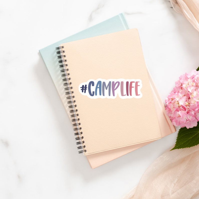 May include: A beige notebook with a black spiral binding and a sticker that says "#CAMPLIFE" in rainbow colors.