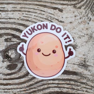 Puede incluir: Una pegatina con una patata de dibujos animados con una cara sonriente y pulgares hacia arriba. El texto "YUKON DO IT!" está en una pancarta circular sobre la patata. La pegatina está sobre una superficie de madera desgastada.