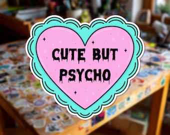 Può includere: Un adesivo a forma di cuore con un bordo smerlato color turchese e un centro rosa chiaro. Il testo "CUTE BUT PSYCHO" è stampato in nero con caratteri in stile gocciolante al centro. Lo sfondo rosa presenta piccoli dettagli a forma di stella.