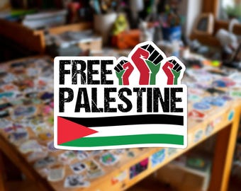 Può includere: Un adesivo fustellato con il testo FREE PALESTINE in un carattere nero dall'aspetto usurato. Sopra il testo ci sono tre pugni alzati in rosso, nero e verde. Sotto il testo c'è una grafica orizzontale della bandiera palestinese con strisce rosse, nere, bianche e verdi.