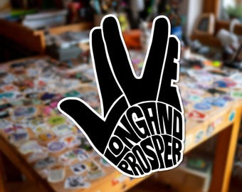 Può includere: Un adesivo in bianco e nero a forma di mano che esegue il saluto vulcaniano. Le dita e il palmo sono riempiti con il testo "LIVE LONG AND PROSPER" integrato nella sagoma nera, caratterizzato da un design del carattere audace e stilizzato.