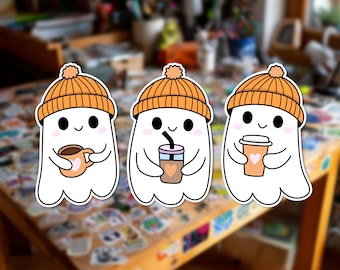 Può includere: Tre adesivi a forma di fantasma bianco, ognuno con un berretto di lana arancione con pon-pon. I fantasmi hanno occhi neri, guance rosa e piccoli sorrisi. Tengono in mano diversi contenitori per bevande: una tazza, un bicchiere con cannuccia.