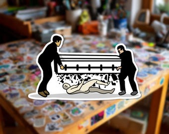 Può includere: Un adesivo fustellato con un'illustrazione grafica di due figure in abito nero che trasportano una bara rettangolare bianca. Sotto la bara, una figura pallida e distesa è raffigurata tra detriti neri sparsi su uno sfondo bianco.