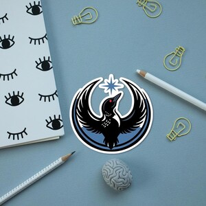May include: A sticker featuring a black bird with outstretched wings, a red eye, and a star, set within a blue and white circular design. The sticker is on a light blue surface with a notepad, pencils, paperclips, and a brain-shaped eraser.