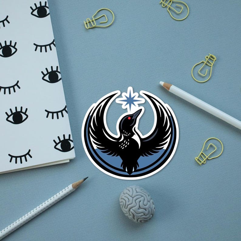 May include: A sticker featuring a black bird with outstretched wings, a red eye, and a star, set within a blue and white circular design. The sticker is on a light blue surface with a notepad, pencils, paperclips, and a brain-shaped eraser.