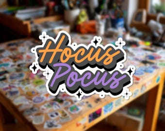 Può includere: Un adesivo fustellato con il testo Hocus Pocus in un carattere stilizzato. La parola Hocus è arancione e la parola Pocus è viola. Entrambe le parole hanno una spessa ombra nera e sono circondate da piccole stelle bianche a quattro punte.