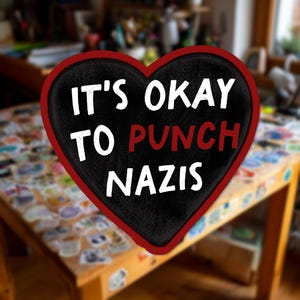 May include: A heart-shaped sign with a black background and a red border. The text "IT'S OKAY TO PUNCH NAZIS" is written in white, with "PUNCH" in red. The sign is likely a statement piece or a form of protest.