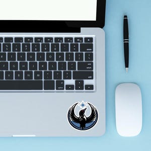 May include: A laptop with a sticker featuring a black bird with outstretched wings, a red eye, and a star. The sticker is on the lower right of the laptop. A black pen and a white mouse are on a light blue surface.