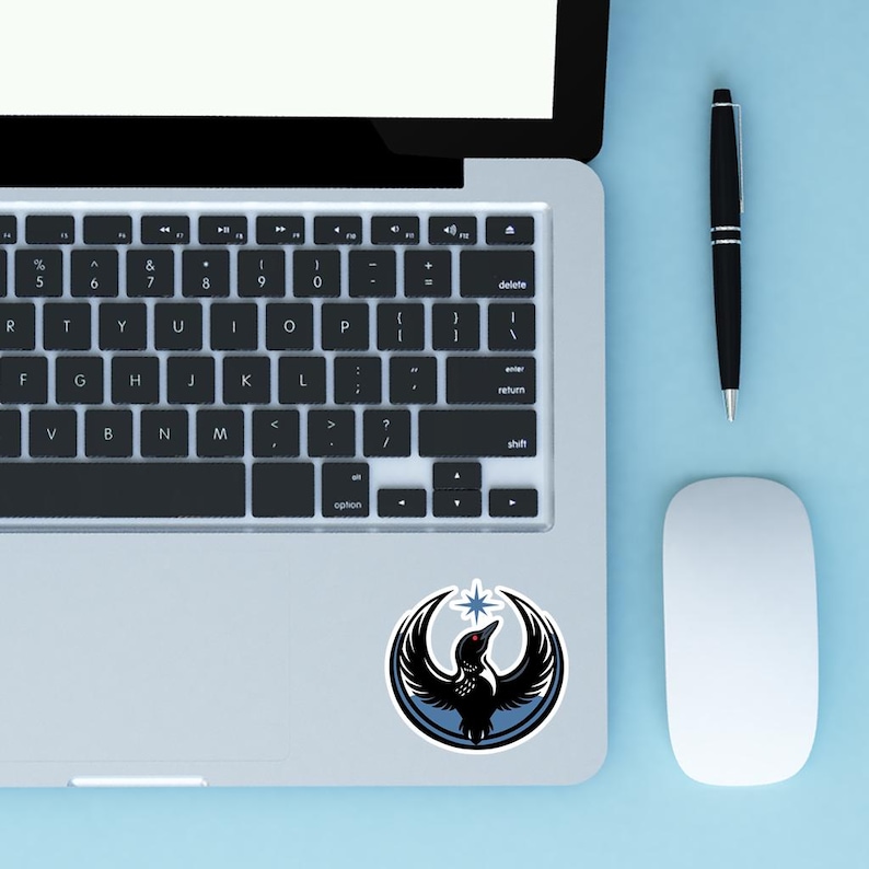 May include: A laptop with a sticker featuring a black bird with outstretched wings, a red eye, and a star. The sticker is on the lower right of the laptop. A black pen and a white mouse are on a light blue surface.