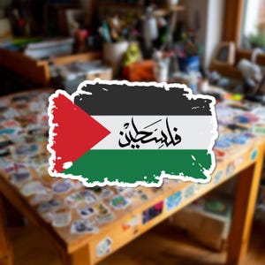 May include: A rectangular sticker featuring the flag of Palestine with a distressed, brush-stroke border. The design includes a black top stripe, a white middle stripe with black Arabic calligraphy, a green bottom stripe, and a red triangle.