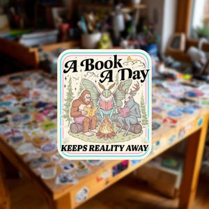 Op de afbeelding: Een rechthoekige sticker met een kleurrijke illustratie van een sasquatch, een motwezen en een figuur met een hertenkop die op boomstammen rond een kampvuur zitten te lezen. De tekst luidt A BOOK A DAY KEEPS REALITY AWAY in zwarte letters.