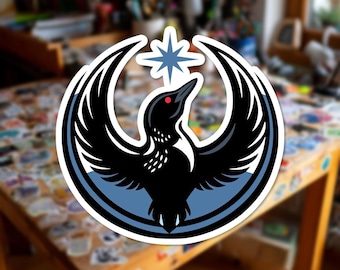 Può includere: Un adesivo raffigurante un uccello nero stilizzato con le ali spiegate, un occhio rosso e una stella blu sopra la testa. L'uccello è posto su uno sfondo circolare blu e bianco. L'adesivo è su un tavolo di legno.