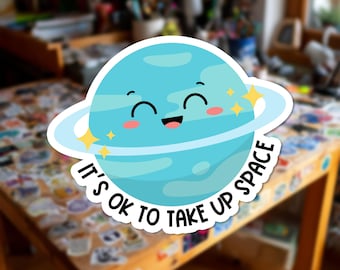 Può includere: Un adesivo circolare raffigurante un pianeta blu chiaro in stile cartone animato con anelli, un volto sorridente e guance rosa. Il testo "IT'S OK TO TAKE UP SPACE" è stampato in lettere maiuscole nere lungo la curva inferiore.