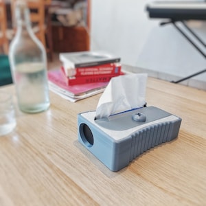May include: A grey and white tissue box, designed to resemble a pencil sharpener, with a single tissue visible. The box sits on a wooden table. A water bottle and books are in the blurred background.