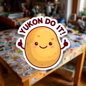 Puede incluir: Una pegatina con una patata de dibujos animados con una cara sonriente, mejillas rosadas y pulgares hacia arriba. La pegatina tiene el texto "YUKON DO IT!" en un diseño circular sobre la patata. El fondo es una mesa de madera cubierta de pegatinas.