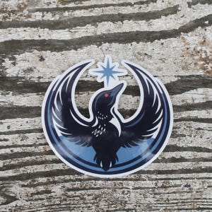 May include: A circular sticker depicting a stylized bird with outstretched wings, in navy and light blue hues. A star is positioned above the bird's head. The sticker is on a weathered, grey wooden surface.