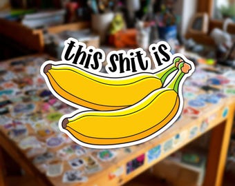 Può includere: Un adesivo fustellato raffigurante due banane giallo brillante con piccioli verdi e punte marroni. Sopra le banane, il testo "this shit is" è scritto in un carattere corsivo nero e audace con un contorno bianco, creando un design.