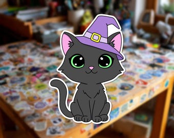 Può includere: Un adesivo fustellato raffigurante un gatto nero con grandi occhi verdi e naso rosa, che indossa un cappello da strega viola chiaro con una fibbia gialla. Il gatto è seduto in posizione frontale con la coda curva verso l'alto sul lato sinistro.