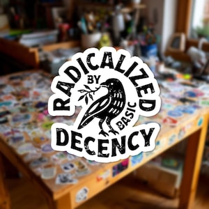 May include: A white sticker with black text and a black bird illustration. The text reads "RADICALIZED BY BASIC DECENCY" in a circular design around the bird holding a sprig. The sticker is on a wooden table covered in other stickers.