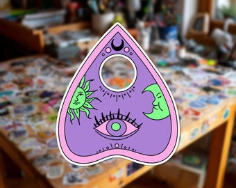Può includere: Un adesivo a forma di planchette triangolare con un centro viola, un sole verde, una luna crescente verde e un grafico a occhio centrale. Il bordo è rosa chiaro con una piccola luna crescente nera in alto e un ritaglio circolare in alto.