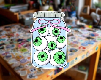 Può includere: Un adesivo raffigurante un barattolo di vetro azzurro riempito con diversi bulbi oculari bianchi con iridi verde brillante e pupille nere. Il barattolo è sigillato con un coperchio grigio e decorato con un nastro rosa chiaro legato al collo.