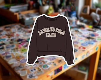 Può includere: Una felpa a maniche lunghe marrone scuro con girocollo e polsini a coste. Sul petto è presente la scritta "ALWAYS COLD CLUB" in un carattere stile college beige chiaro con un contorno marrone scuro, centrata sulla parte anteriore.