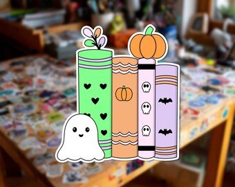 Può includere: Un adesivo fustellato raffigurante una pila di quattro libri in verde chiaro, arancione, bianco e lavanda. Il libro verde ha cuori neri, quello arancione una zucca, quello bianco teschi e quello lavanda pipistrelli neri. Un fantasma è davanti.