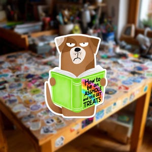 May include: A die-cut sticker featuring a brown dog with a grumpy expression holding an open green book. The book cover displays the text How to be an ASSHOLE and still GET TREATS in black lettering against a rainbow-colored background.
