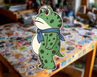Può includere: Un adesivo fustellato raffigurante una rana dei cartoni animati con pelle verde, macchie verde scuro e pancia color crema. La rana è in piedi di profilo, rivolta verso sinistra, e indossa una semplice sciarpa blu annodata attorno al collo.